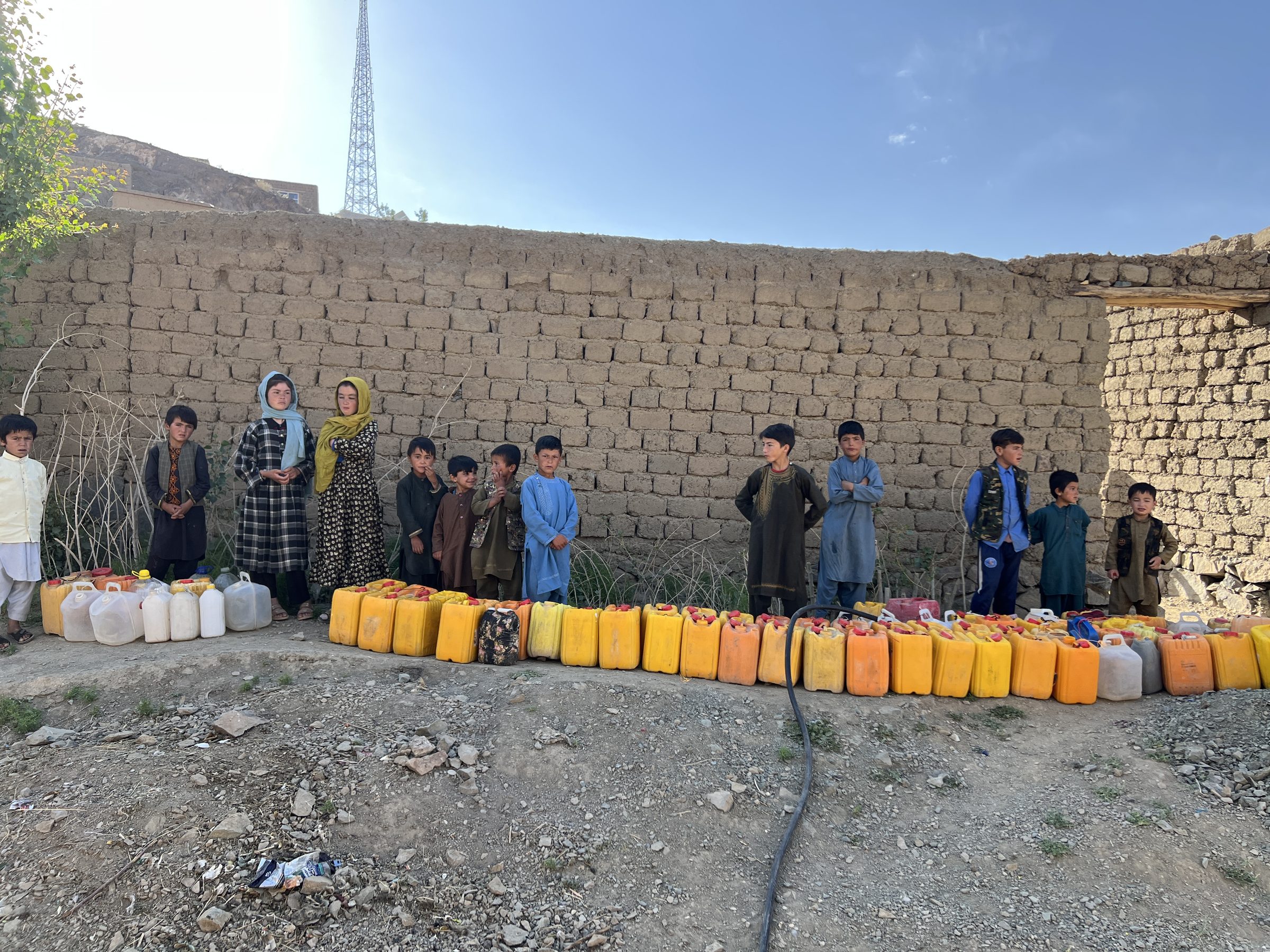 A long line of children beside an even longer row of water jerrycans along a mud-brick wall in Ghor